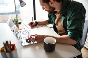 un homme prenant des notes manuscrites tout en étant sur son ordinateur portable